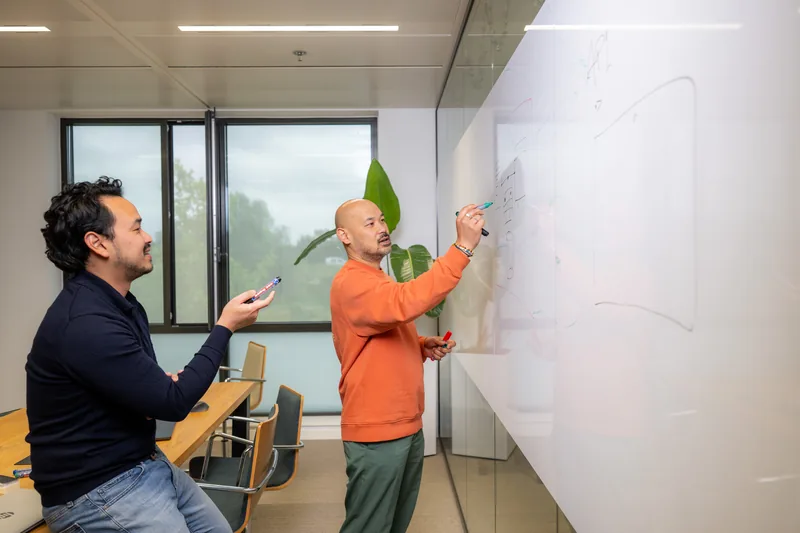 Two colleagues working together at a whiteboard, one sketches while the other looks on with a phone in hand